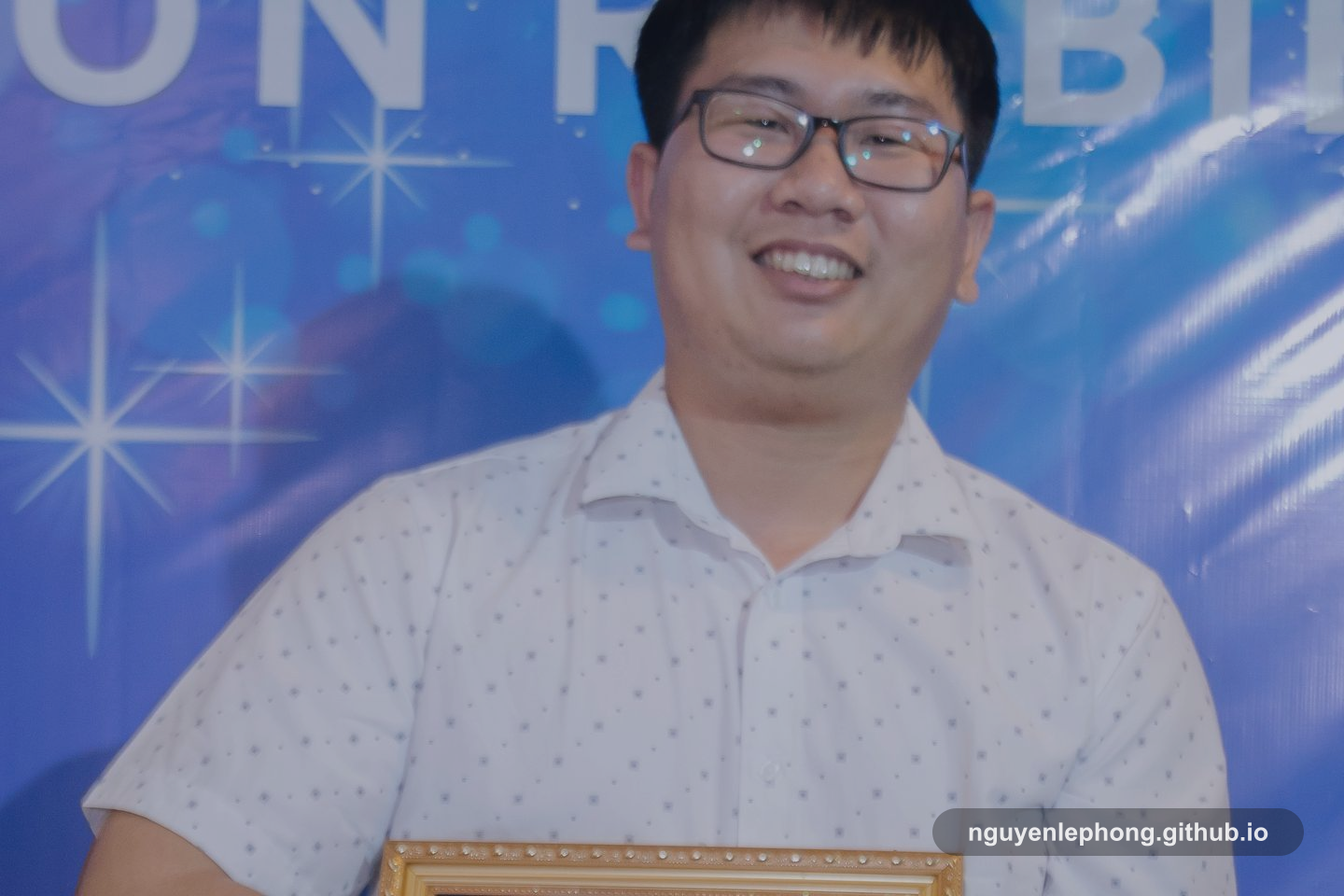 Nguyen Le Phong mỉm cười sau một khoảnh khắc được ghi nhận công khai, phía sau là nền sự kiện được làm mờ để cảm giác được công nhận nổi lên rõ hơn phần banner.