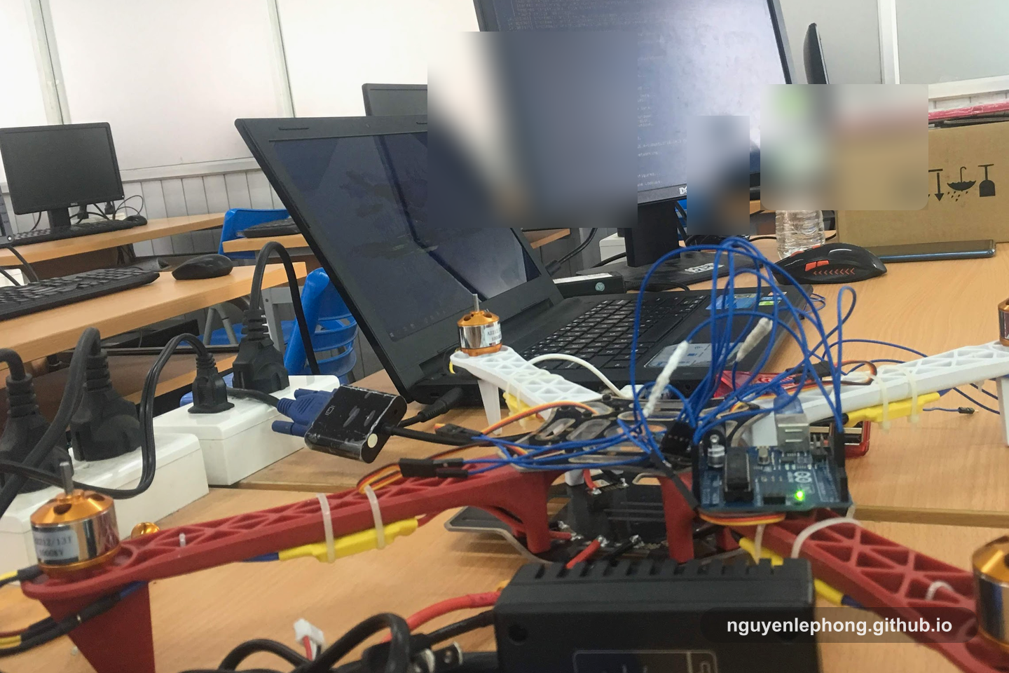 Một mẫu drone nằm cạnh laptop trong phòng lab đại học, với dây nối, dụng cụ và thiết bị thử nghiệm còn trải trên bàn.