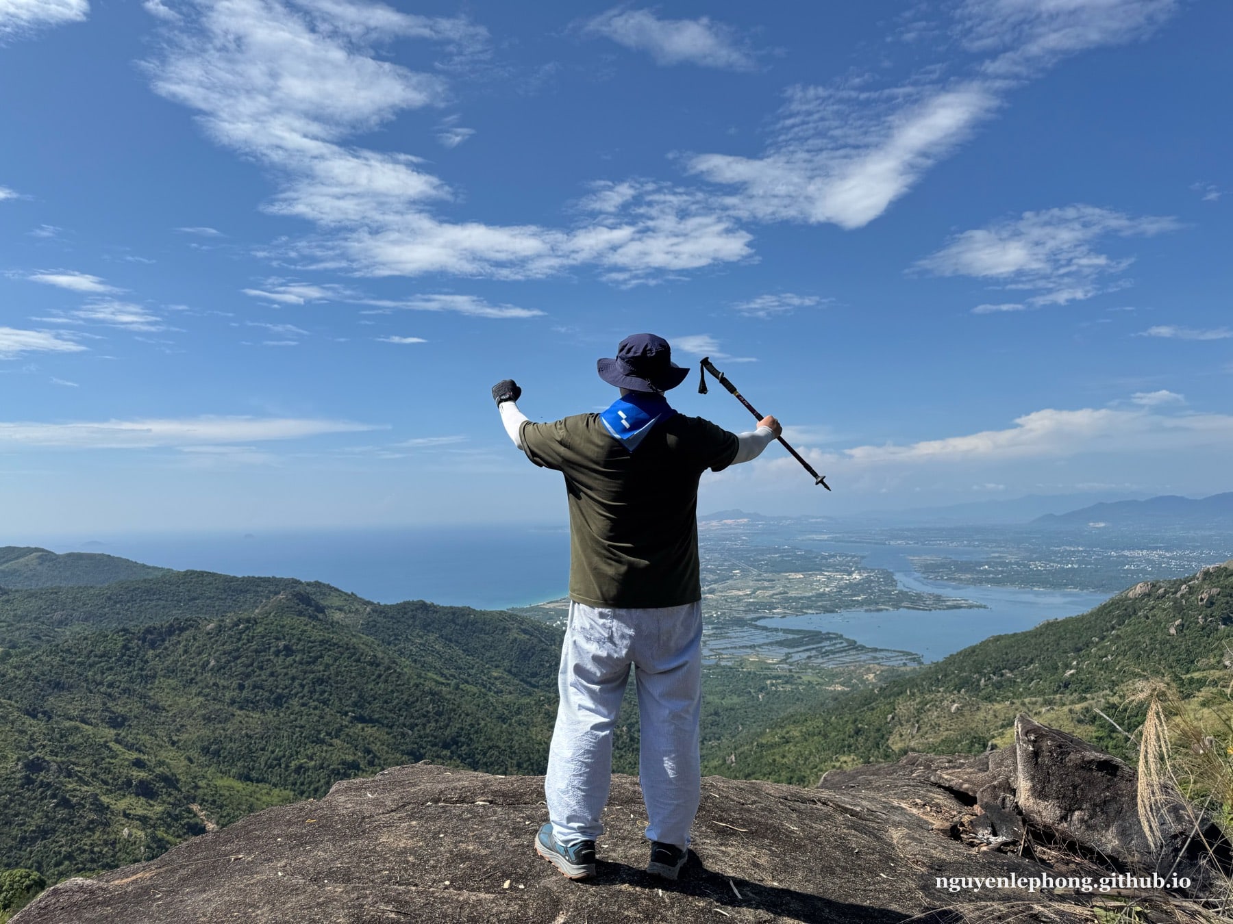 Tác giả đứng quay lưng trên tảng đá đỉnh, dang tay và cầm gậy trek phía trên vịnh biển và các dãy núi