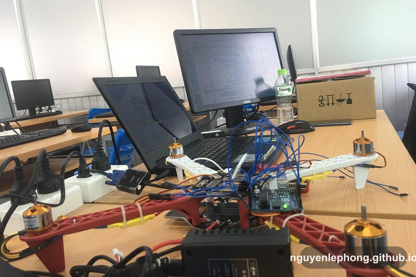Khung drone còn lộ dây và linh kiện nằm cạnh laptop và dàn màn hình trong lớp học, trước khi interface cuối cùng che mất các phần chuyển động bên dưới.