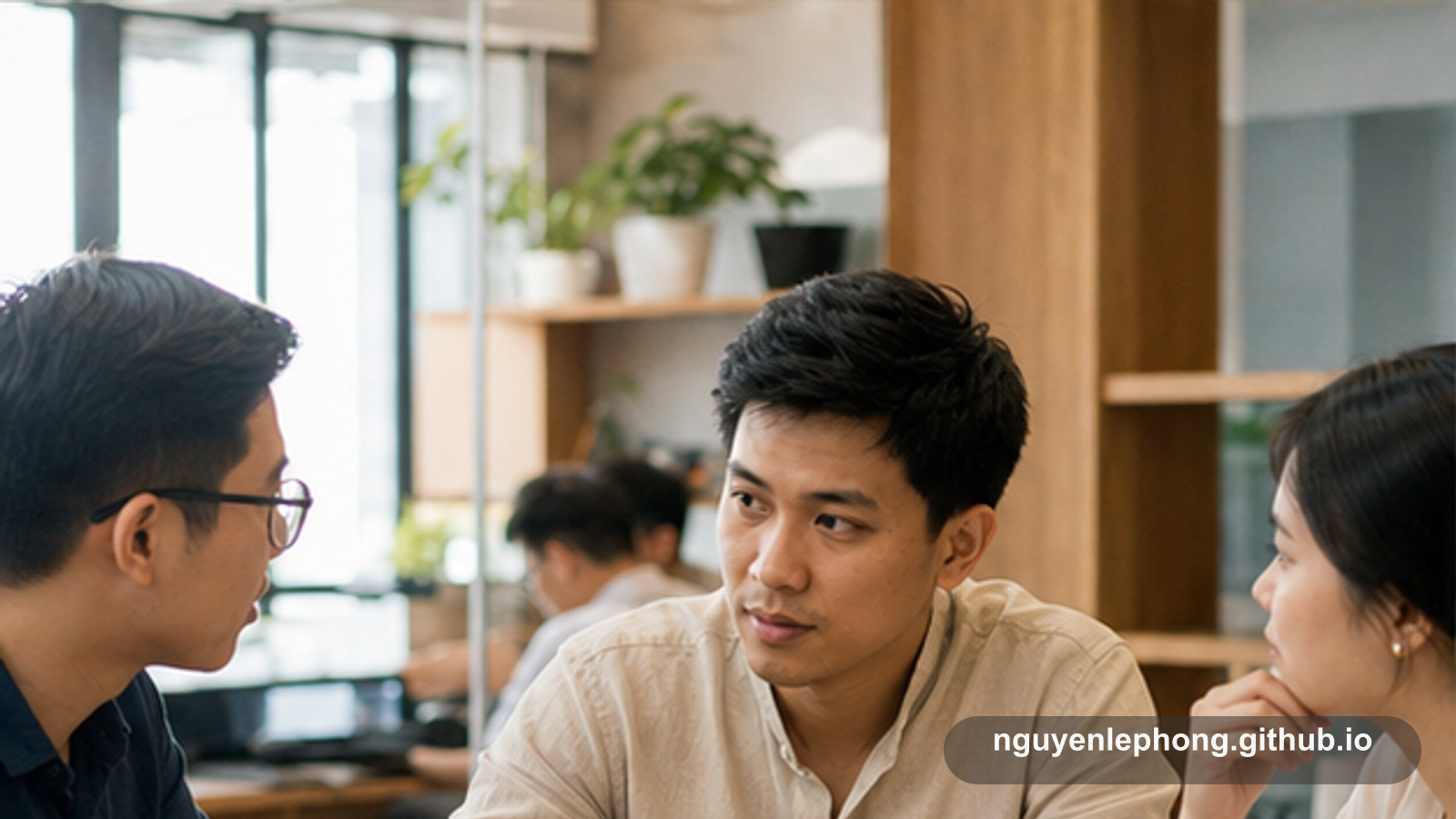 Nguyen Le Phong nghiêng người lắng nghe hai đồng đội trong một không gian làm việc ấm áp, đủ chậm để hiểu bối cảnh của họ trước khi phản hồi.