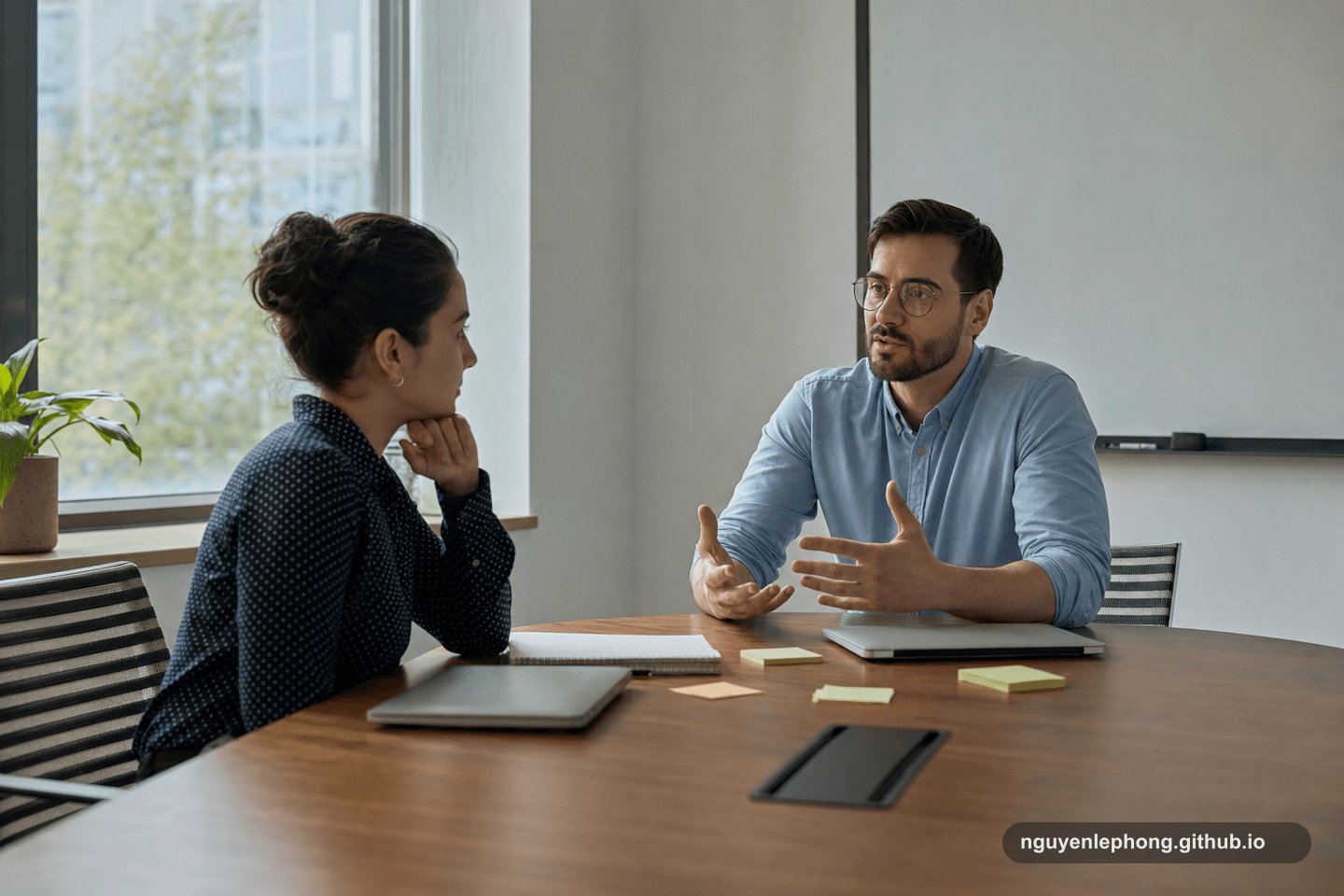 Two coworkers sit in a quiet meeting room after a hard review, speaking calmly across closed laptops and plain notes.