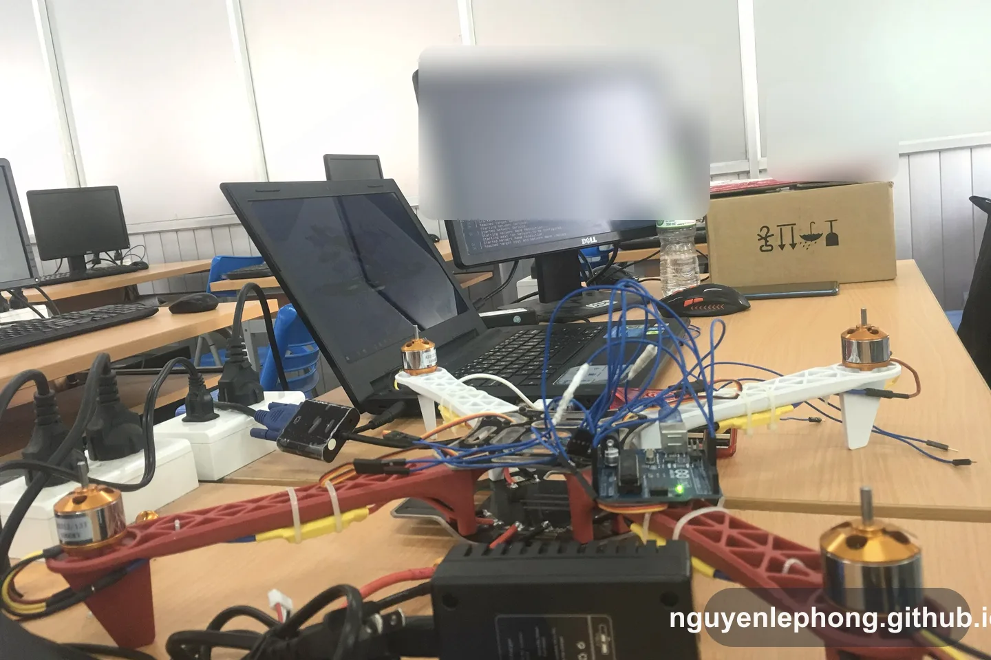 Một prototype phần cứng, laptop và màn hình trên bàn làm việc, gợi việc ý tưởng phần mềm vẫn phải đi qua thiết bị thật và ràng buộc thật.