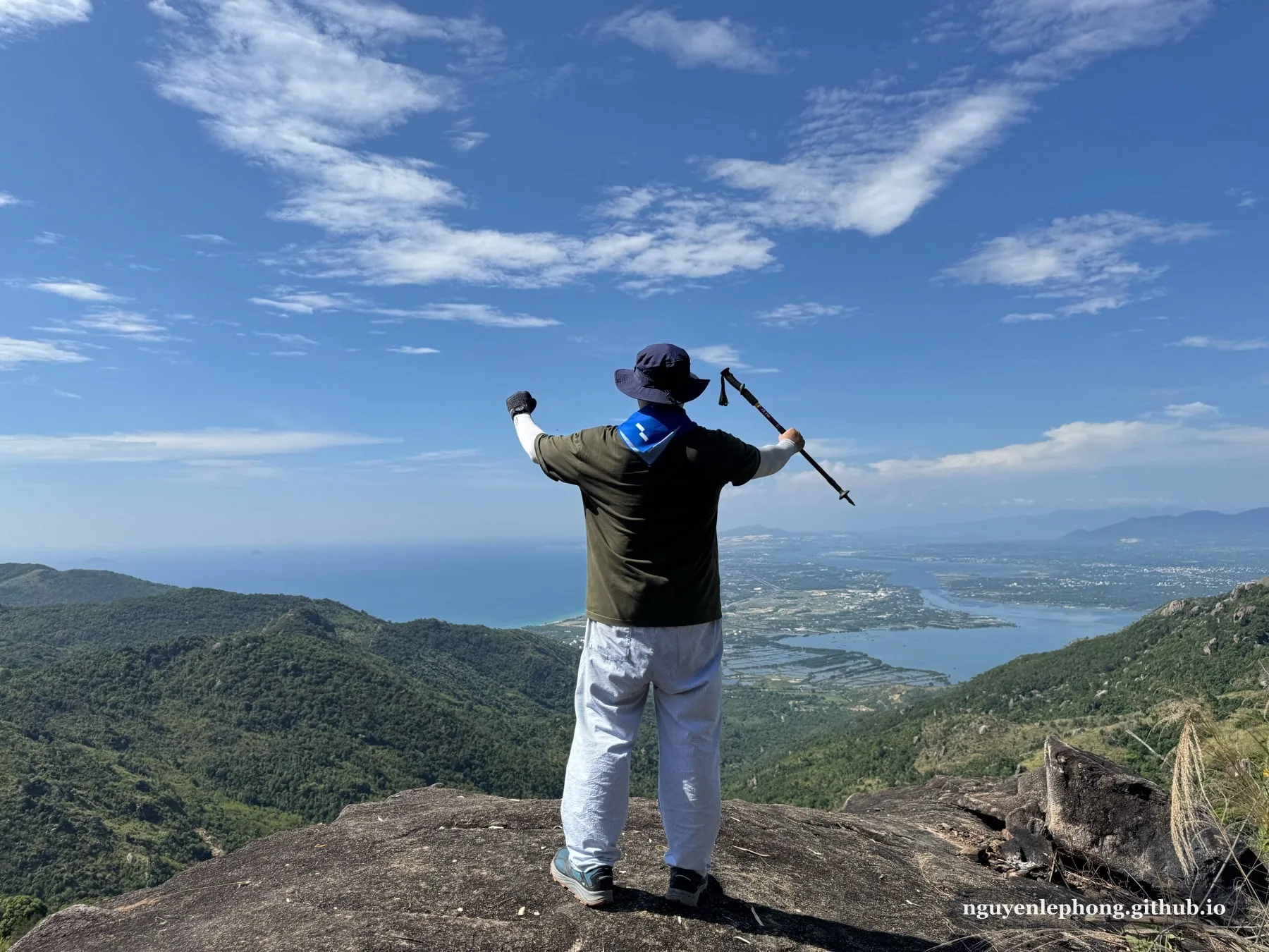 Tác giả đứng quay lưng trên tảng đá đỉnh, dang tay và cầm gậy trek phía trên vịnh biển và các dãy núi