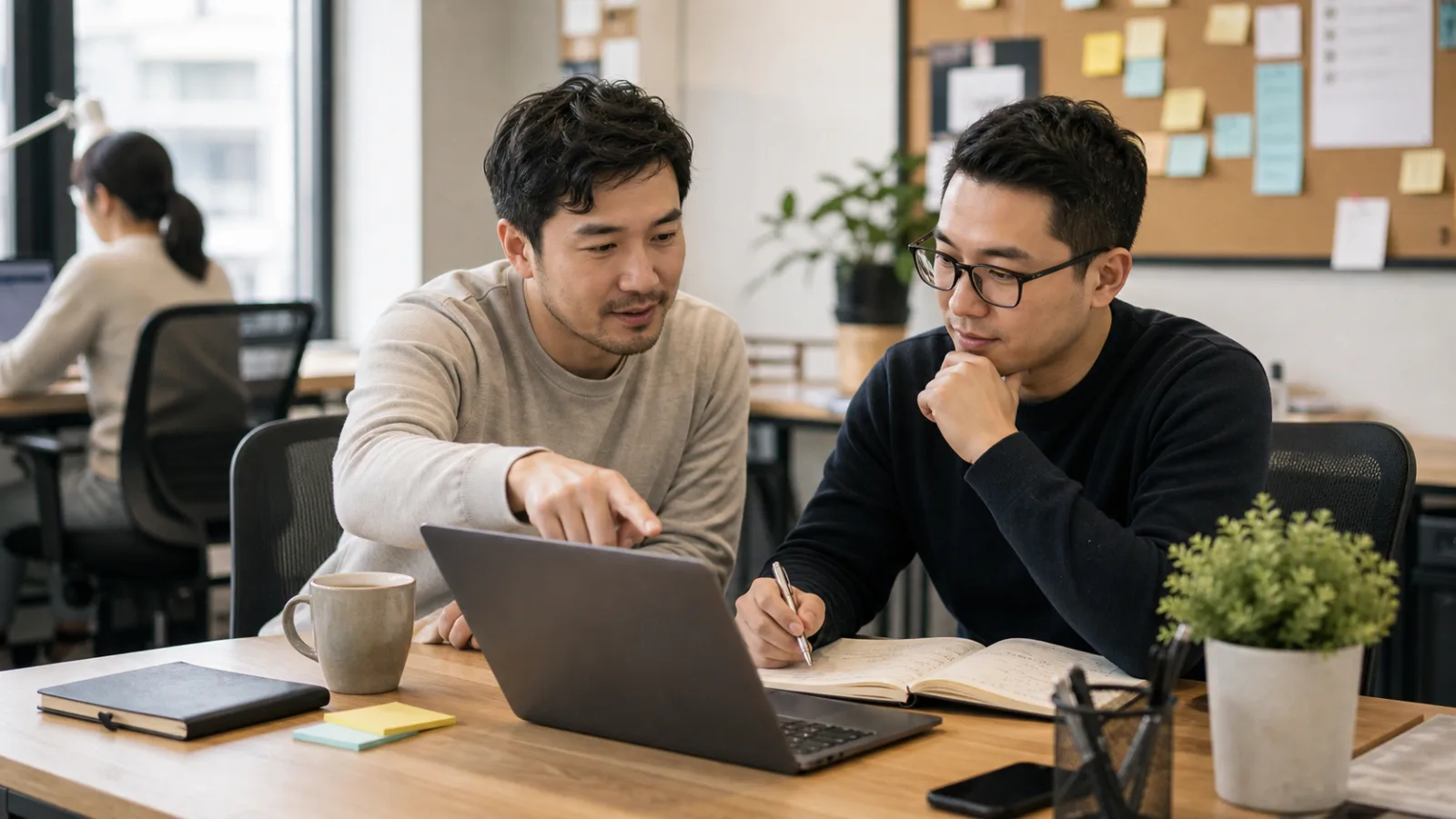 Hai đồng đội ngồi ở bàn làm việc, cùng nhìn vào laptop trong khi một người giải thích vấn đề và người còn lại ghi chú.