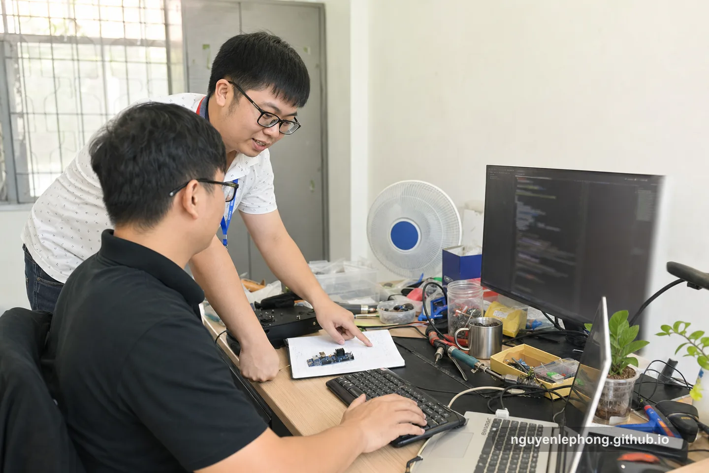 A Vietnamese engineering lead leans beside a teammate's desk, pointing at one technical detail while the teammate keeps control of the keyboard.
