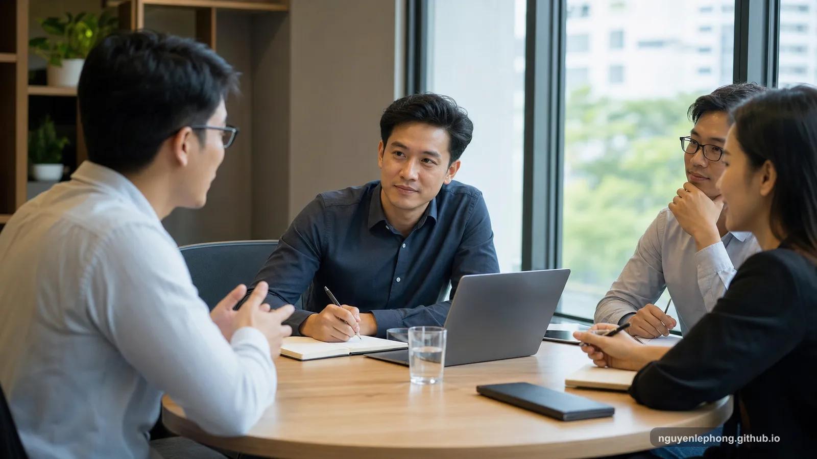 Nguyen Le Phong sits at a round meeting table and listens carefully while another teammate speaks, keeping the room open instead of pulling attention toward himself.